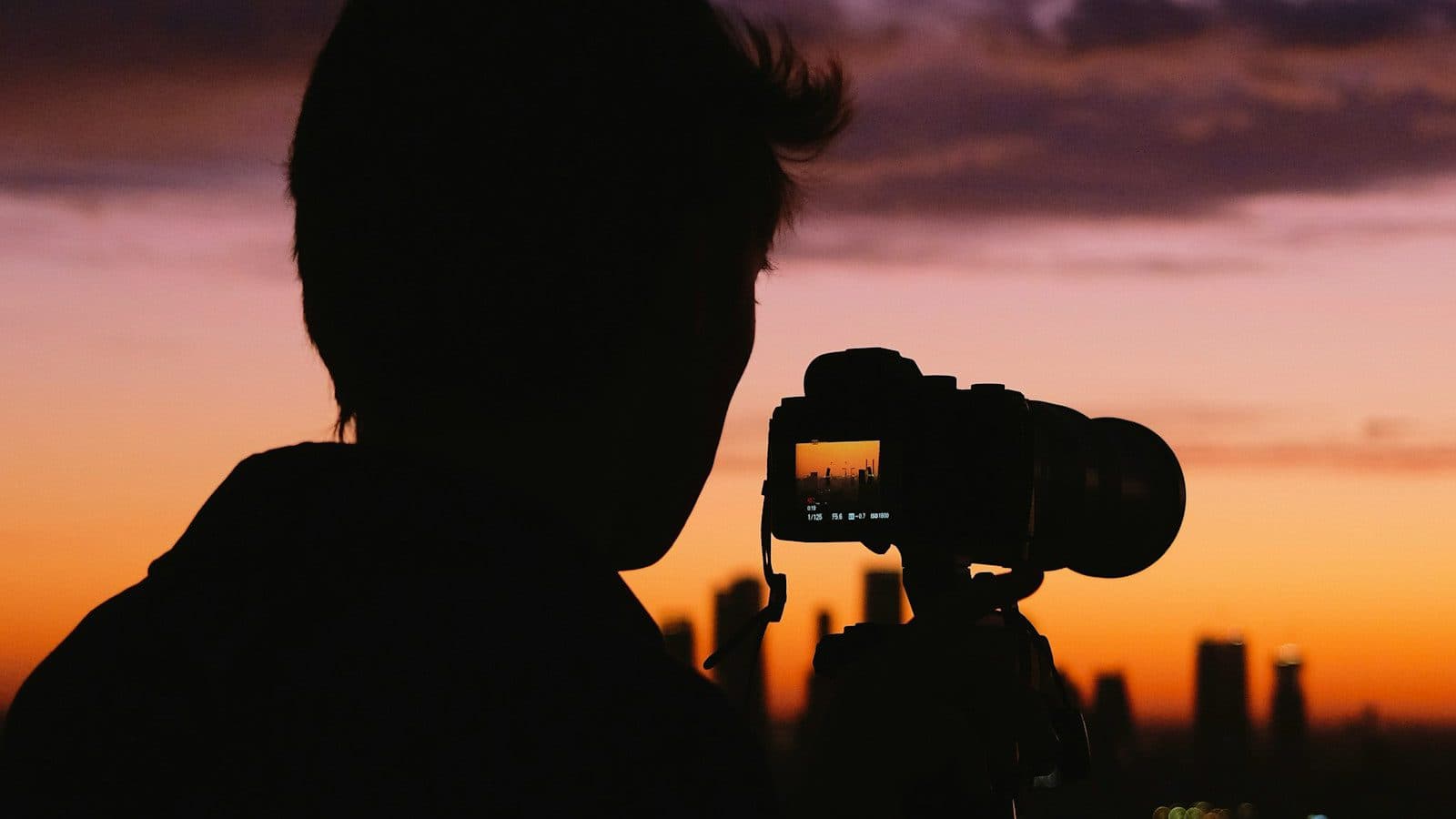 Silhouetted photographer framing a city skyline on a tripod-mounted camera at sunset